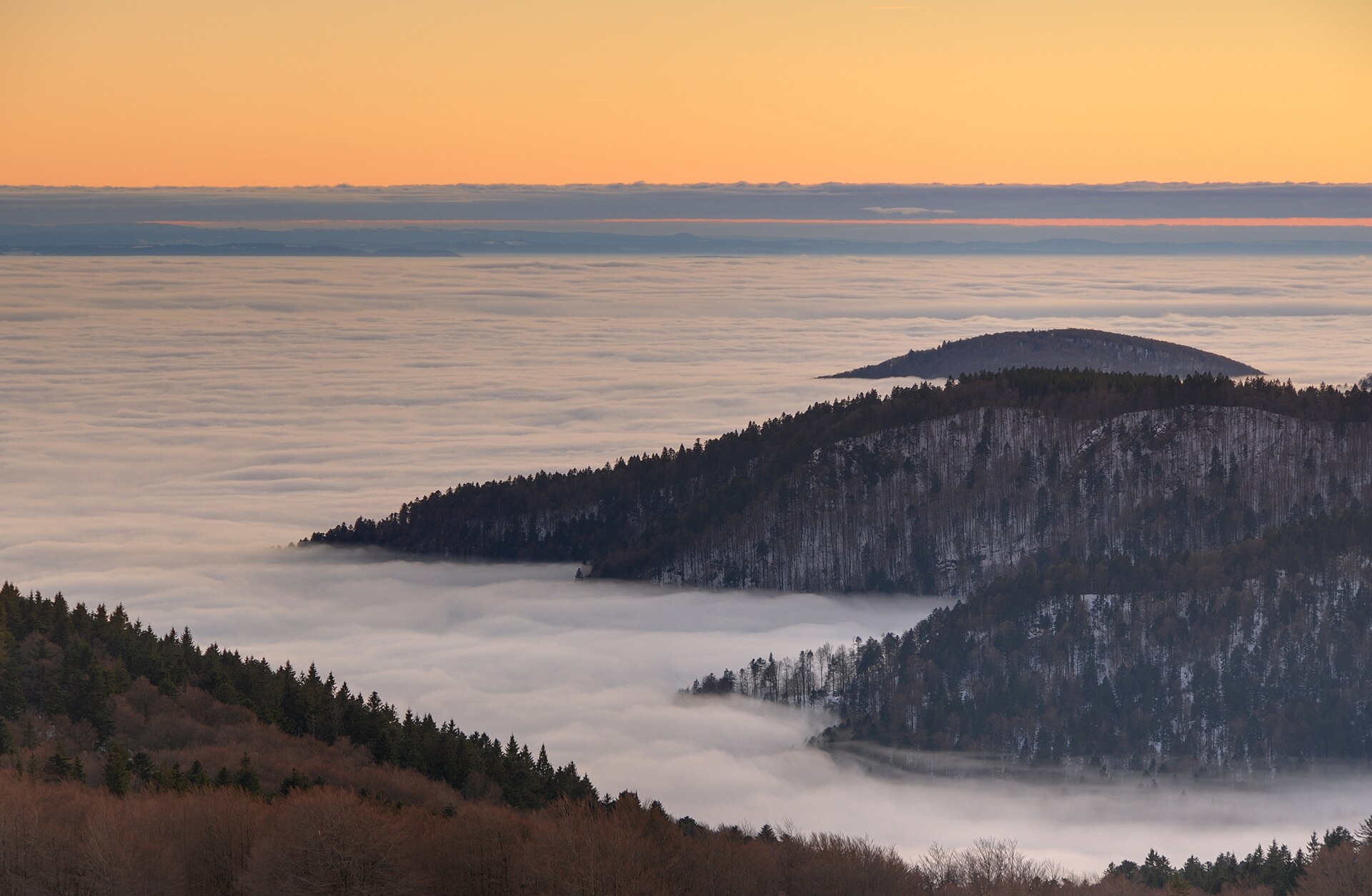 A sea of fog filling a valley below dark wooded hills at sunset